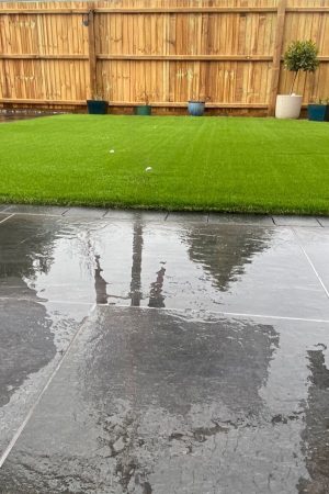 Wet dark slate patio tiles reflecting light beside a vibrant artificial turf lawn and tall wooden fence with decorative potted plants.